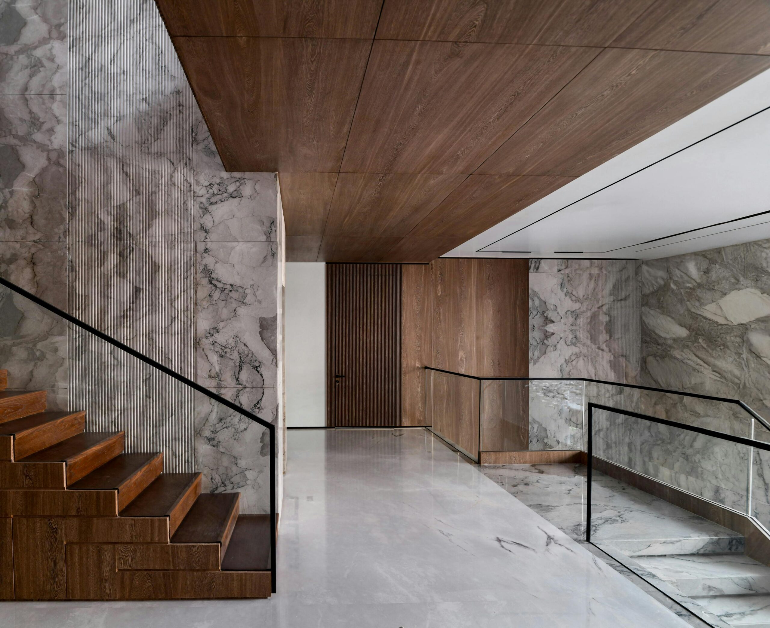 Sleek modern hallway with marble walls, wooden ceiling and stairs, and glass railings.
